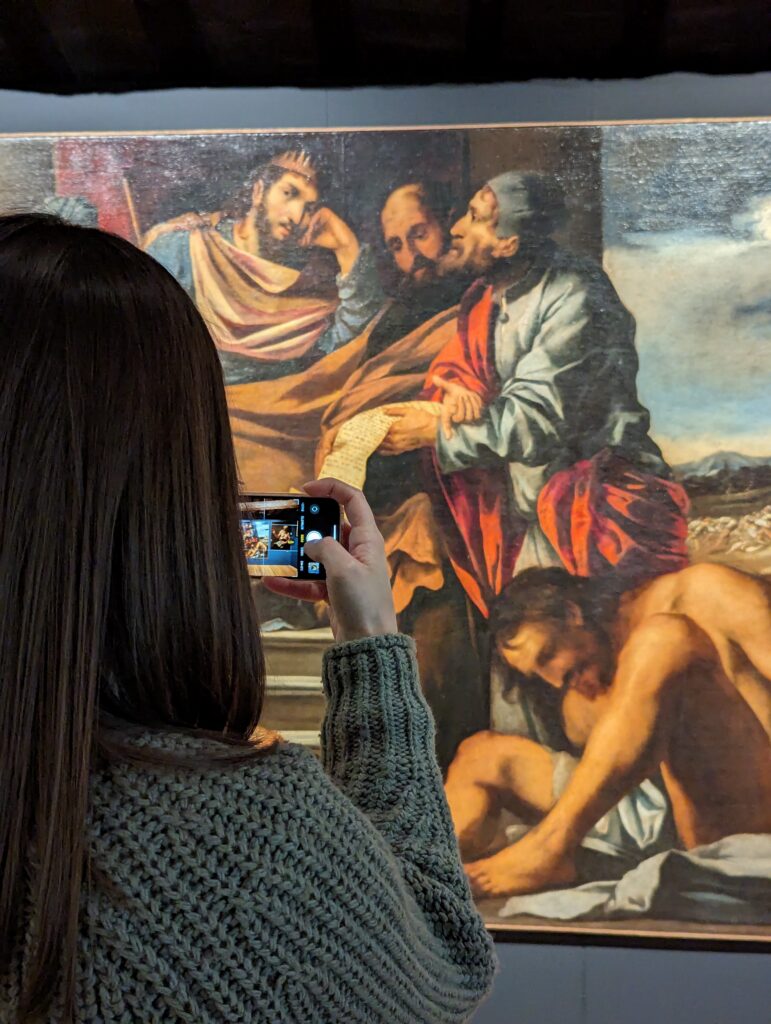 Una ragazza di spalle sta fotografando un dipinto antico.